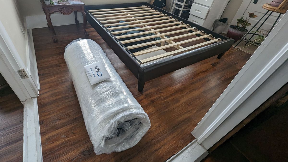 Puffy Lux Hybrid mattress vacuum packed and rolled in plastic, next to a bed frame
