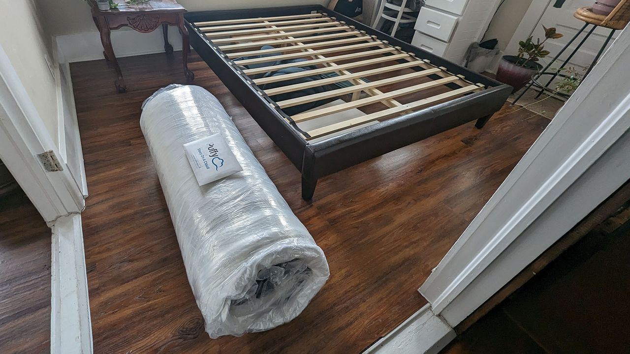 Puffy Lux Hybrid mattress vacuum packed and rolled in plastic, next to a bed frame
