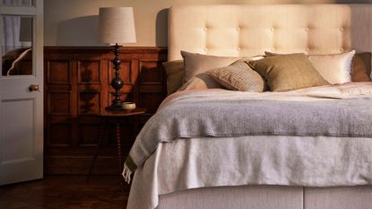Neutral bedroom space with a cream upholstered bed to the right, with neutral linen bedding, and wood panelling behind the bed. To the right of the bed is a small circular wooden beside table, with a dark wood lamp with white lampshade, and a white open door beside it.