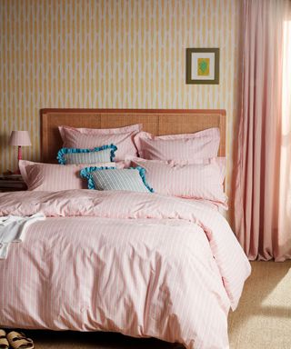 A baby pink bed with pink and blue pillows and a white throw on it on it, yellow patterned wallpaper, and dusky pink curtain drapes