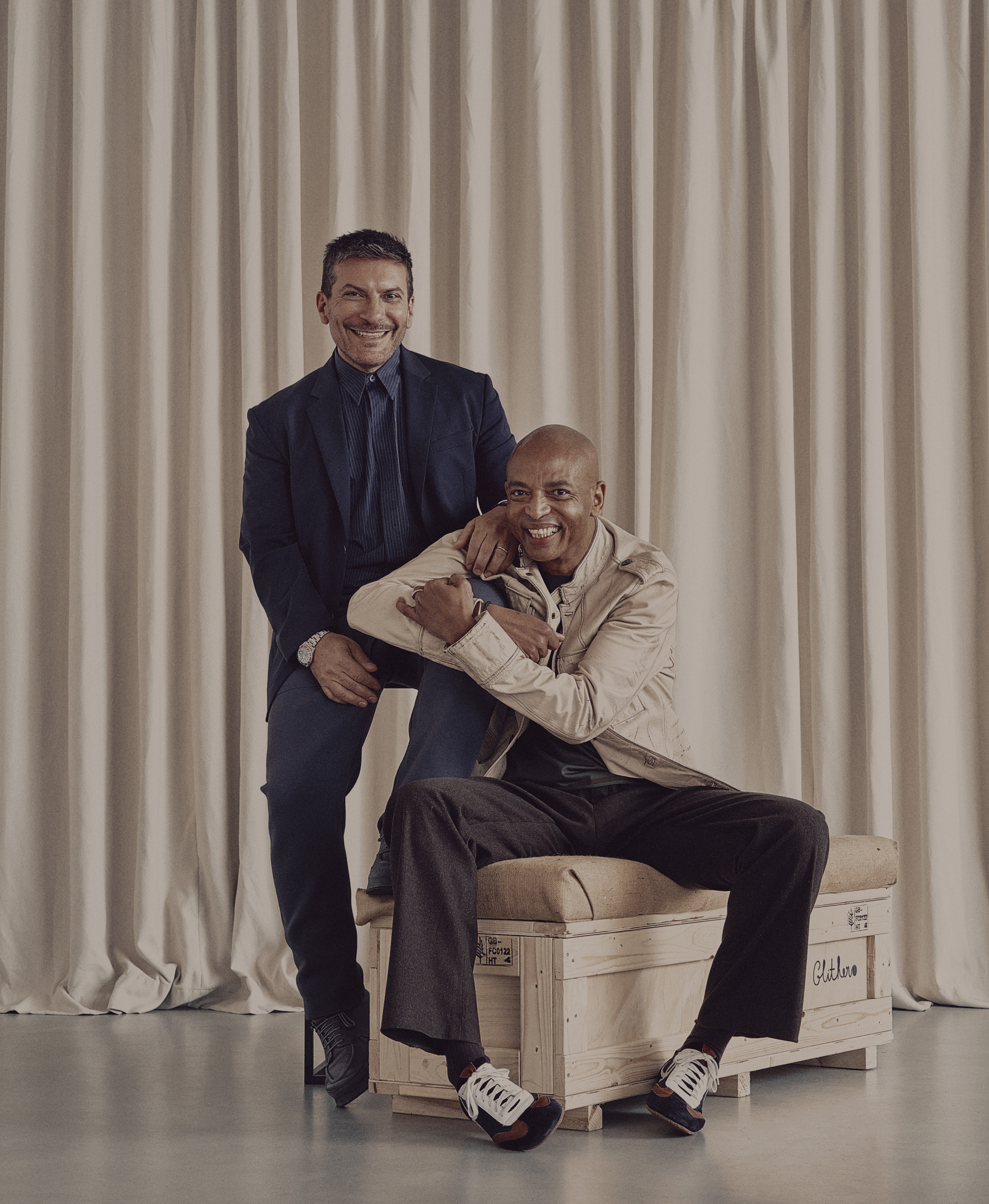 Two man dressed in smart-casual clothing, the first one in a dark blue cotton suit, the other one in a beige trenchcoat, dark gray trousers, and brown and black sneakers, pose for the camera in a bare room decorated with beige curtains and a wood and fabric stool.
