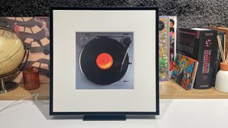 Samsung Music Frame wireless speaker on white surface in front of busy shelf showing turntable picture in frame