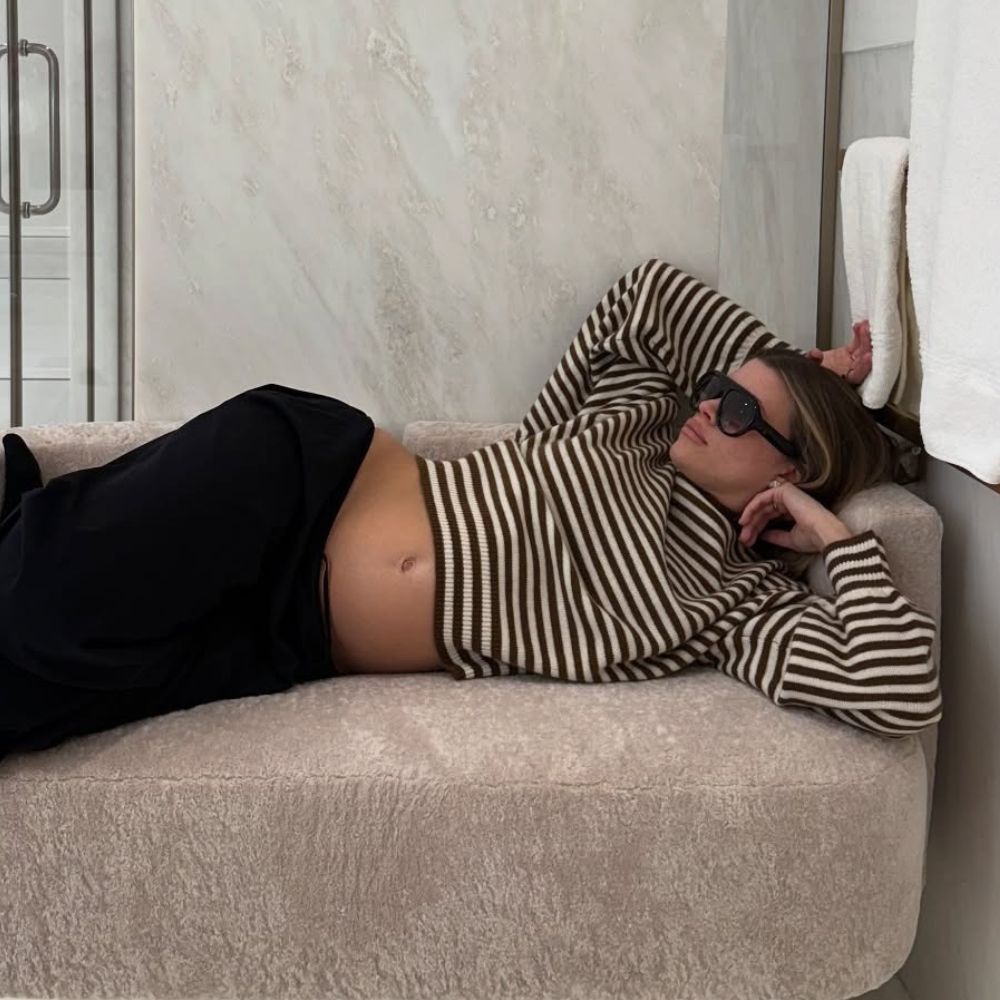 Sofia Richie Grainge lies on a sofa wearing a black and white striped knit with track pants and black sunglasses.