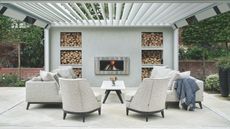 A large concrete patio with white upholstered chairs and sofa with white table, large white awning and central log burner with recessed wood storage