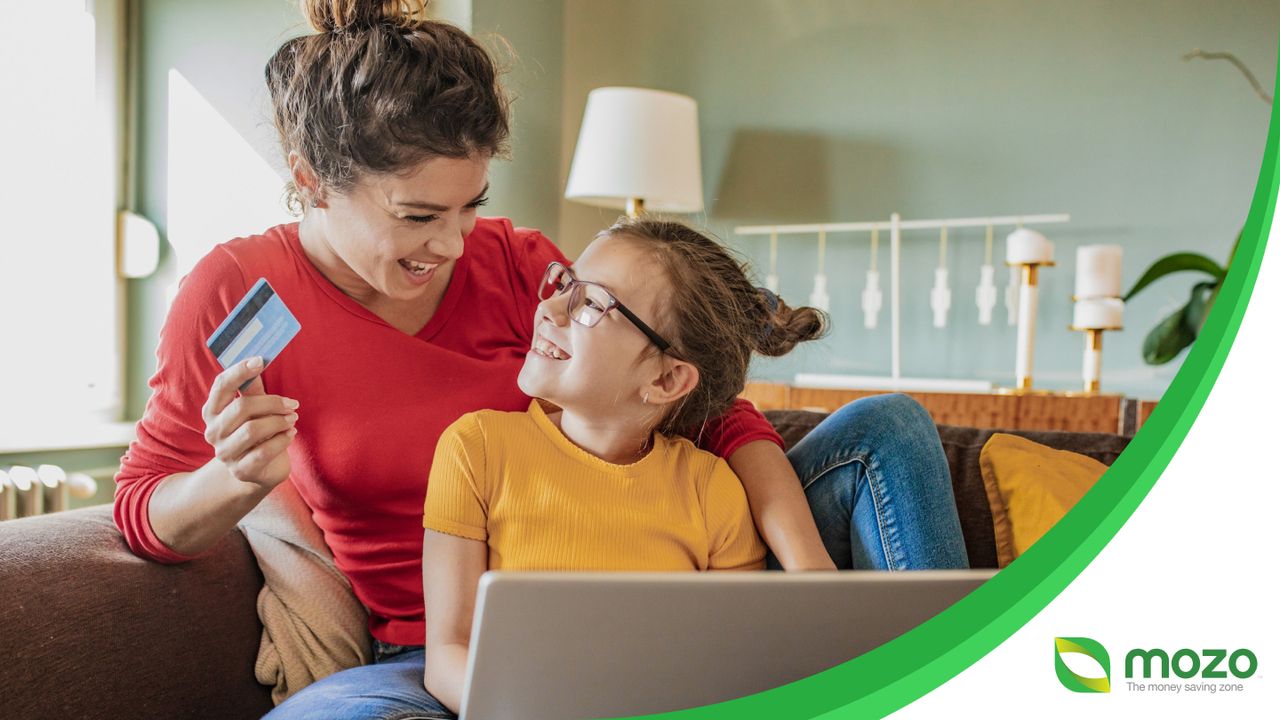 A mother and daughter sitting on the couch using a laptop to shop online. The mother is holding a bank card in one hand.