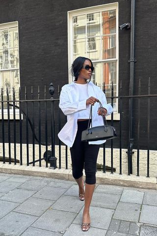 how to make a button-down shirt look more expensive is shown in a photo of a woman standing on the sidewalk in London wearing a white button-down shirt over a white tank top with black capri pants, black sunglasses, a black shoulder bag, and black mules