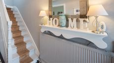 A general view of a hallway with black and white checked tiled floor, bare wood staircase, and radiator beneath ornate shelf with the letters spelling the word HOME