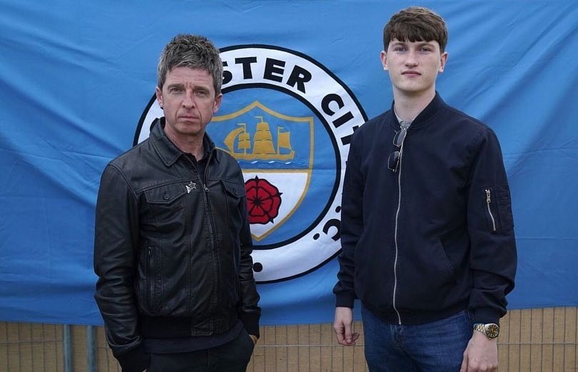 Noel Gallagher and Andrew Cushin stand in front of a Manchester City flag, highlighting their connection to football culture.