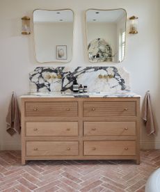 A white bathroom with terracotta floor tiles, and wooden vanity with a marble countertop, and gold vanity mirrors