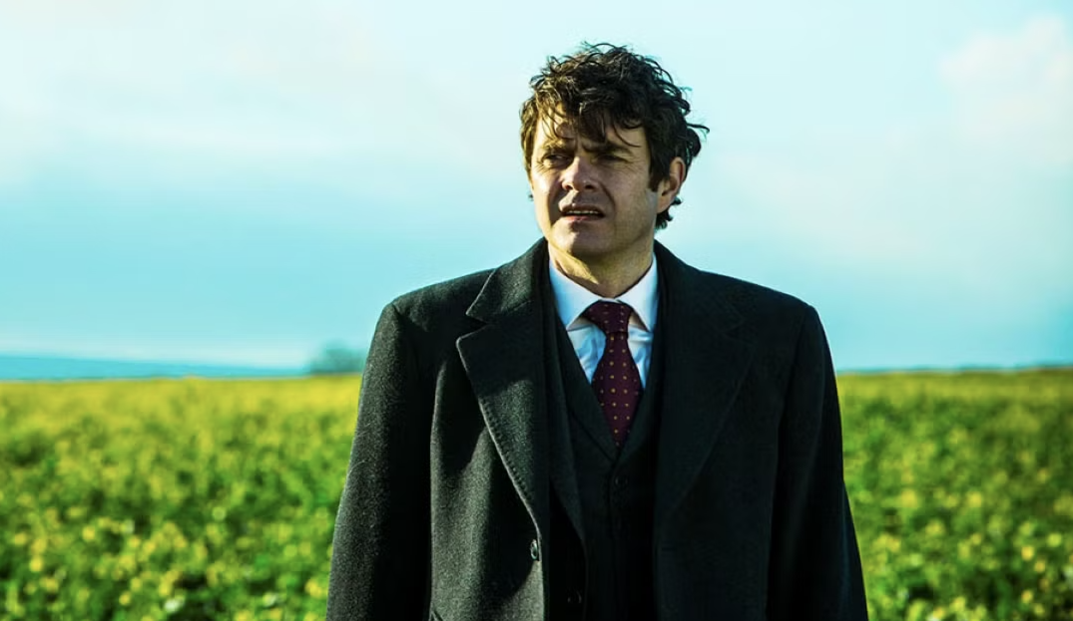 Paul Higgins wears a suit in a Utopia and stands in front of a field