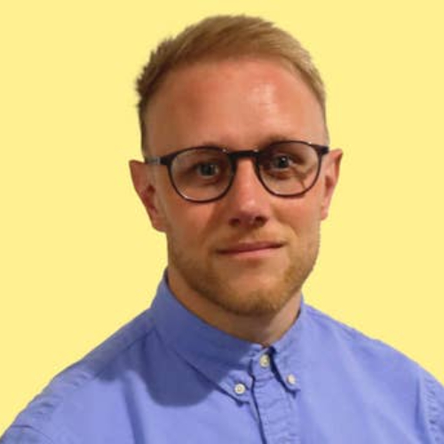 male wearing blue shirt with short hair and black framed glasses stood against yellow background