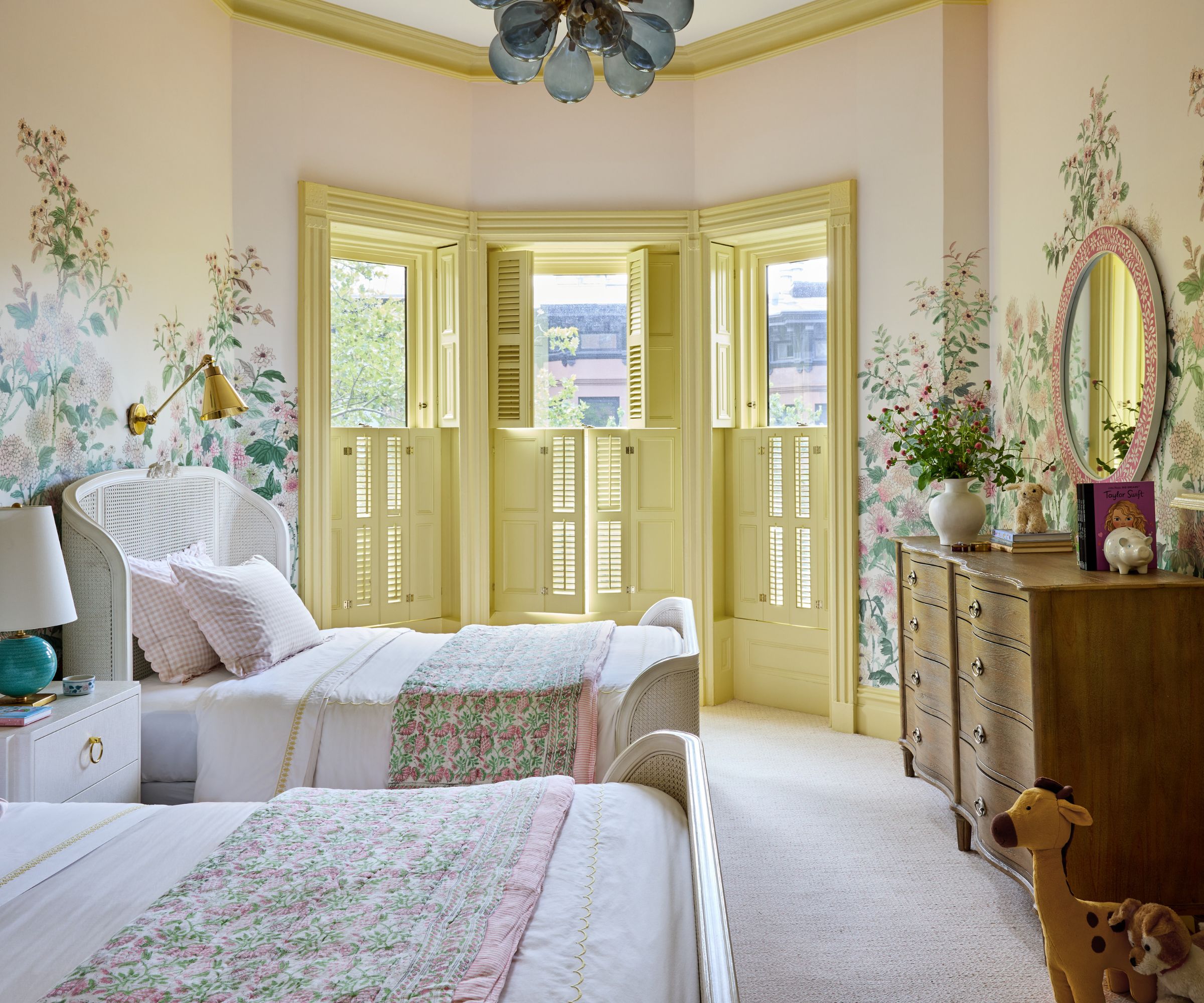 A large bedroom with traditional decor, twin beds, a butter yellow bay window, and a floral wall mural.