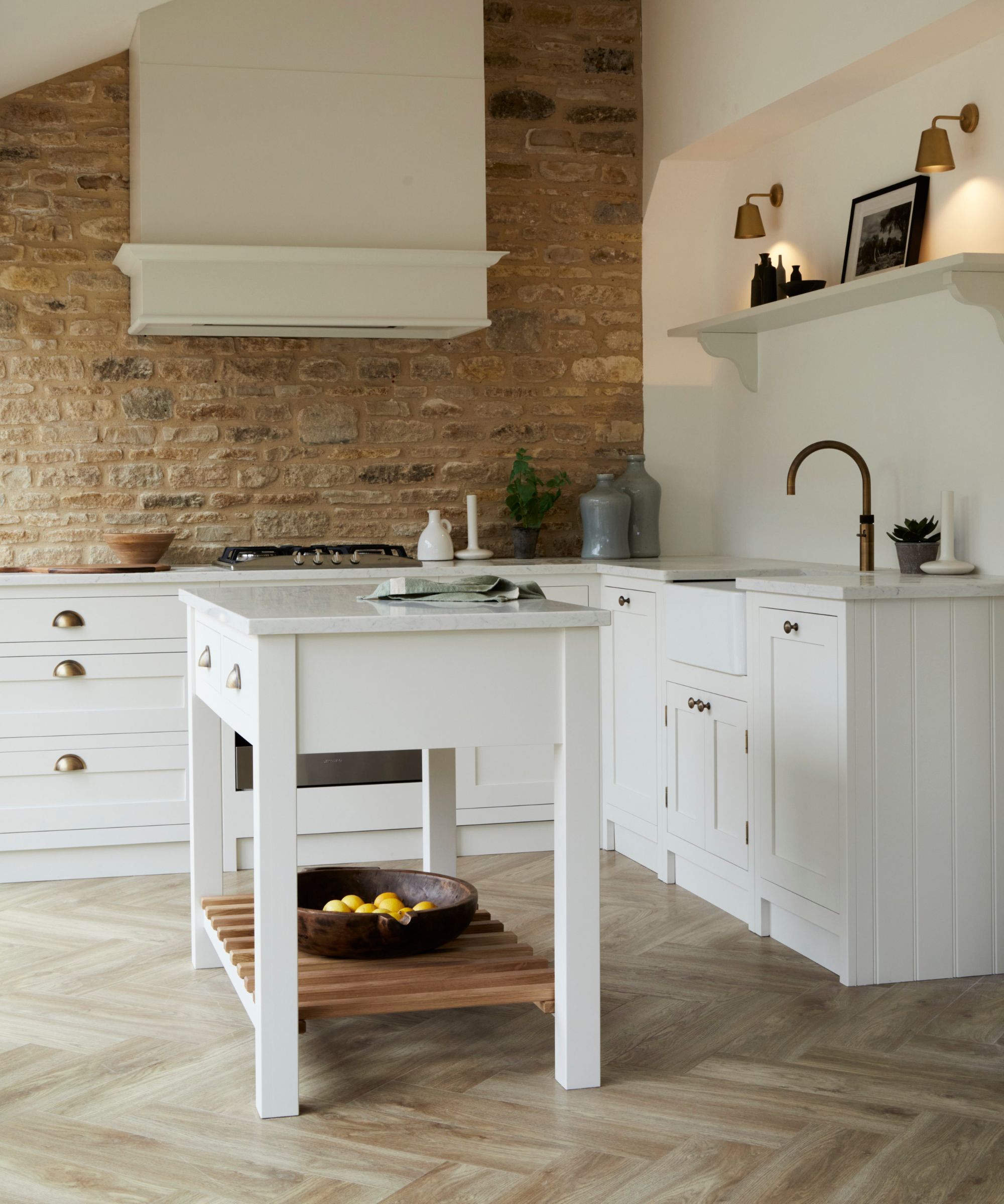 A classic white kitchen with a small freestanding island, wooden worktop, brass handles and exposed brick for a warm, farmhouse feel.