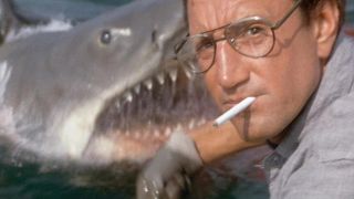 Still from a Jaws movie. Here we see a man wearing glasses and a cigarette in his mouth staring directly at the camera. In the background, you can see a giant great white shark coming out of the water.