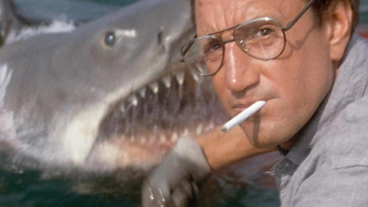 Standbild aus einem Jaws-Film.  Hier sehen wir einen Mann mit Brille und einer Zigarette im Mund, der direkt in die Kamera starrt.  Im Hintergrund sieht man einen riesigen Weißen Hai, der aus dem Wasser kommt.