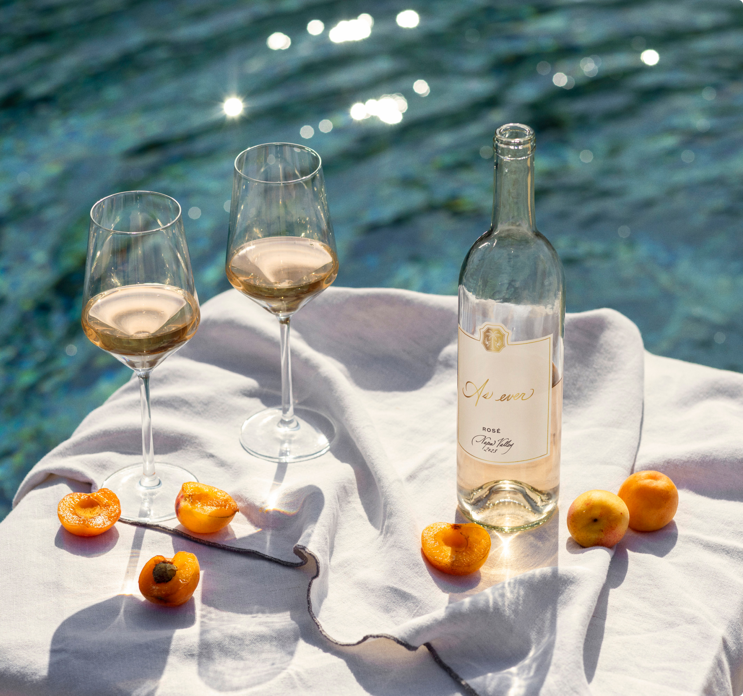 A bottle and two glasses of As ever rosé surrounded by halved apricots photographed on a white cloth in front of a pool