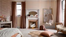 A kids bedroom with pastel walls, floor-length orangey-pink curtains an abstract wallaper on one wall, an unused fireplace in the middle with an oval mirror above, a hanging swing chair, a daybed and a bed and desk with a jute flower-shaped rug and cushion on the floor