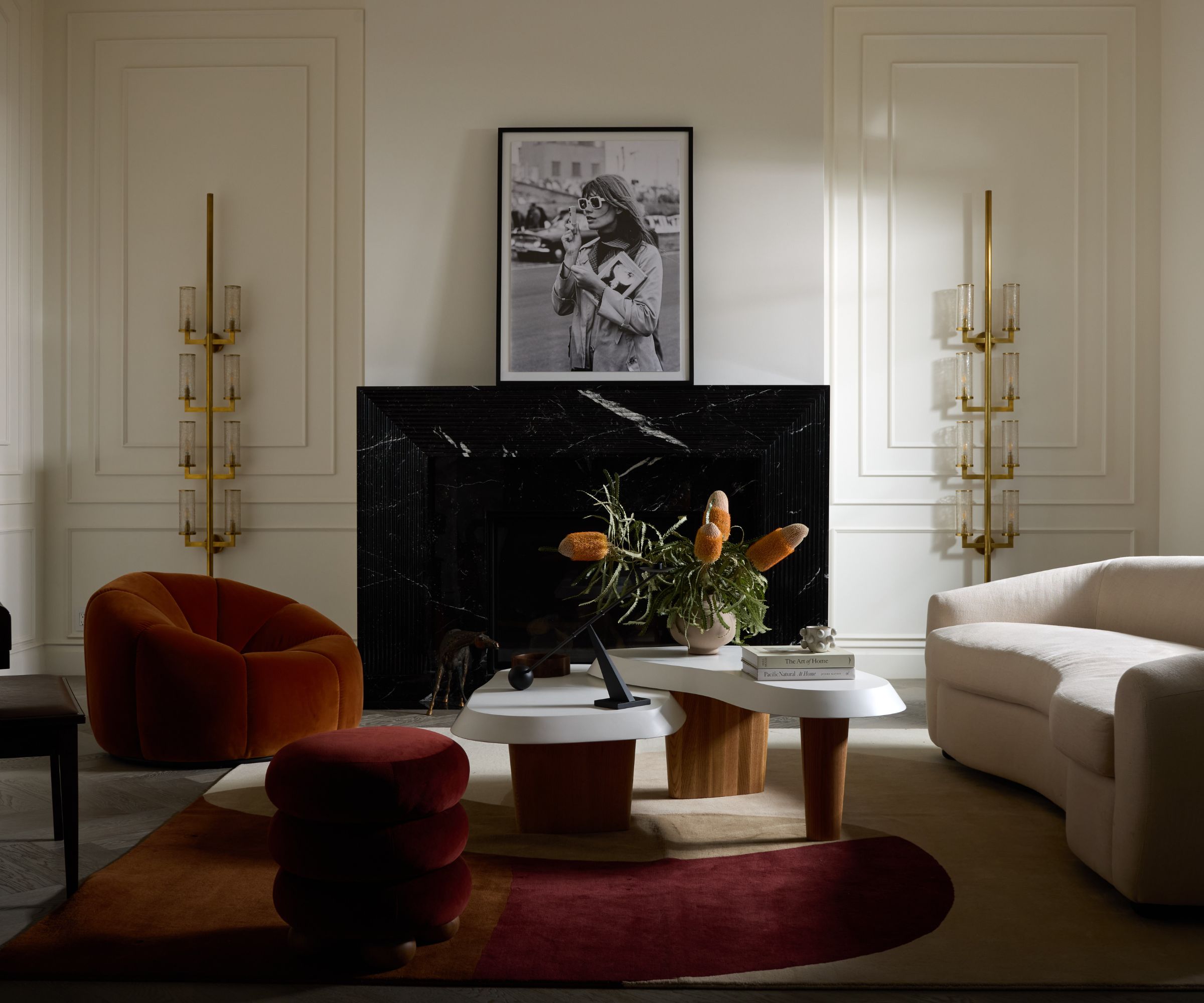 Close up of furniture in front of a black marble fireplace, mantel decorated with artwork, sculptural metallic wall light to the left hand side, geometric rug underneath furniture