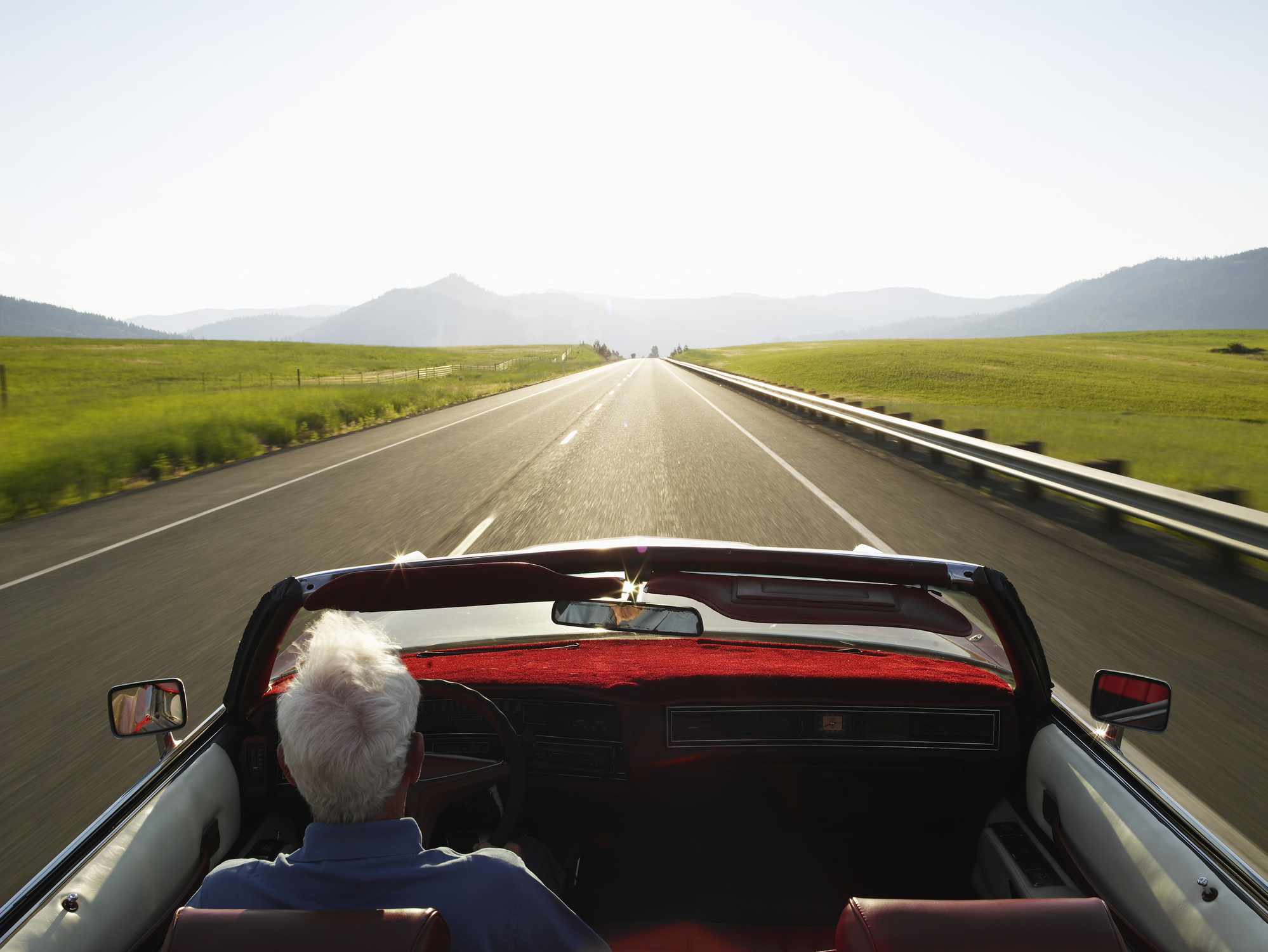 A senior man drives a convertible car down a long, open road.