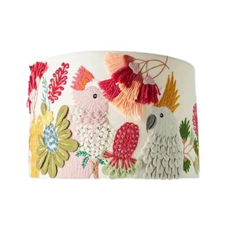 A white lampshade with cockatoo embroidered birds on it with light pink and blue bords, dark pink and coral tassels, and green flowers