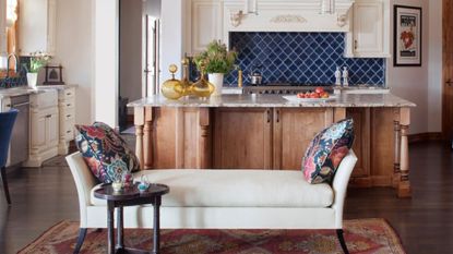 A kitchen with a white storage bench with a red rug underneath, natural and white wooden kitchen cabinets, and a marble kitchen island with yellow vases and strawberries on top