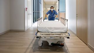 a nurse in blue scrubs and a white face masks pushes an empty gurney down a hospital hallway and towards the camera