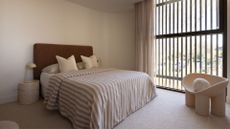 A white bedroom with wood panelled windows, a white curved seat with a circular cushion, a large bed with a brown headboard and a striped bedscape, alongside a stone table with a lamp