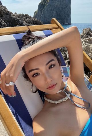 Shell jewelry trend shown on a woman lounging in a beach chair wearing a shell necklace with gold hoops and a blue swimsuit.