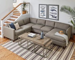 A small modern coastal farmhouse living room with l-shaped grey sofa, grey wooden coffee table, framed wall art, striped area rug and staircase in background