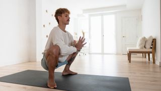 A man performs a yoga garland pose in a white room on a mat. He is in a deep squat position, with his toes pointing outwards and his hand in prayer position, elbows pushing against his knees. We see a floor-length window behind him and two low wooden chairs.