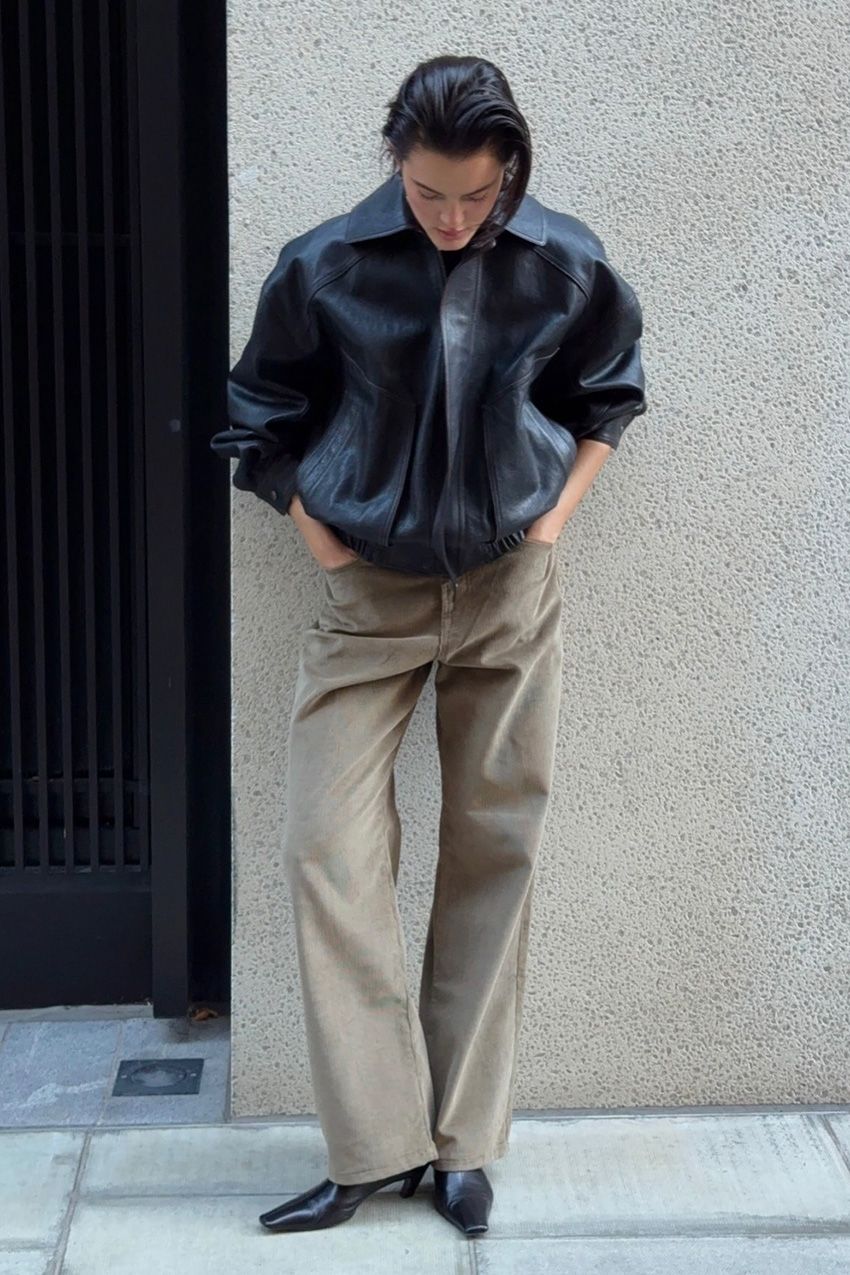 A woman standing outside leaning against a concrete wall wearing an oversize black leather bomber jacket with light brown wide-leg pants and black pointed toe ankle boots.