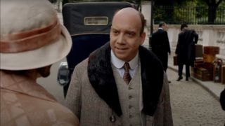 Paul Giamatti half smiles as he stands in a period costume in front of Laura Carmichael in Downton Abbey.