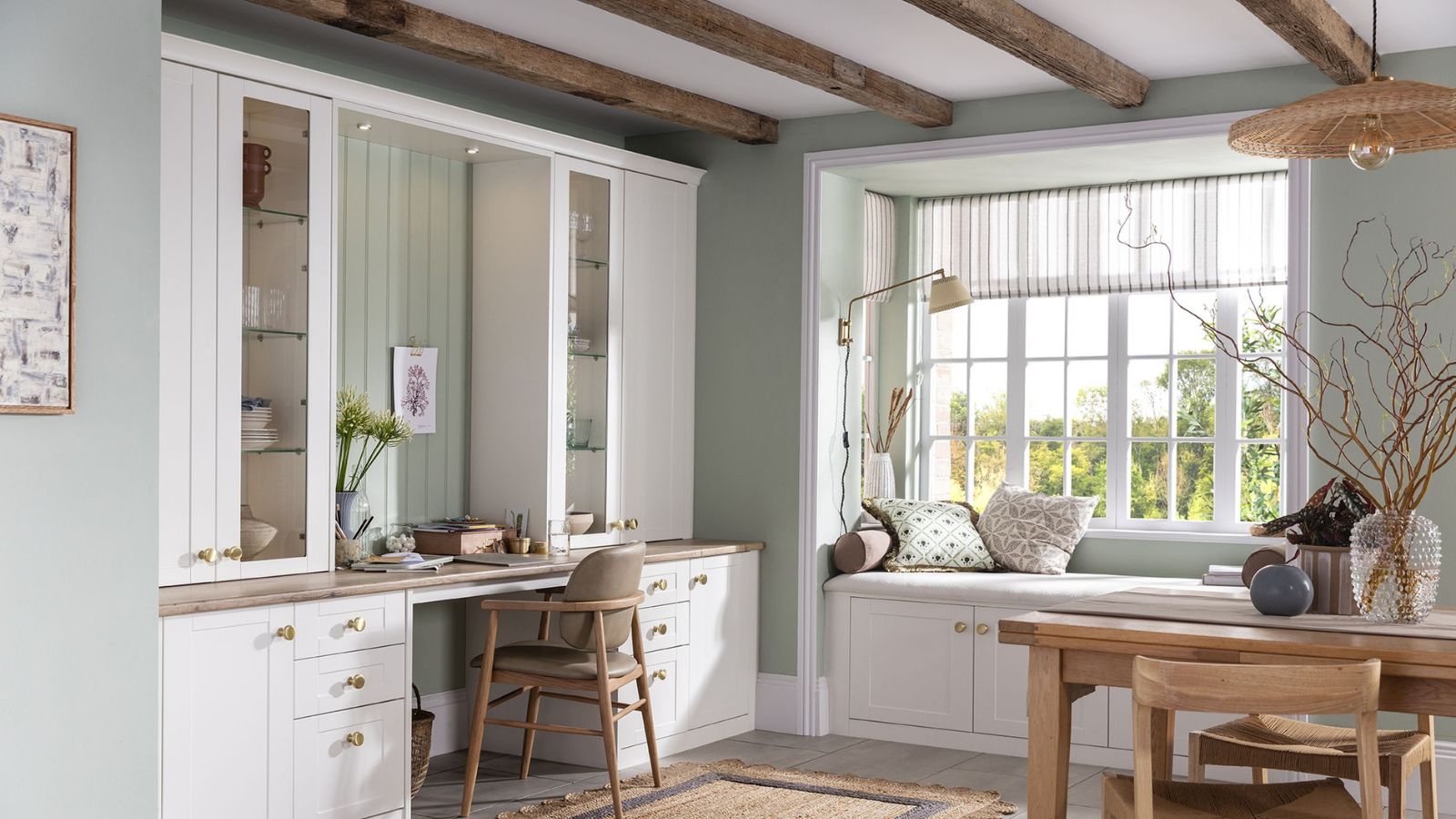 A soft green dining room is shown with built-in white cabinetry forming a small desk area, complete with a wooden chair and glass-fronted cupboards. To the right, a large bay window with a cushioned window seat brings in natural light, framed by striped blinds and scattered patterned pillows.