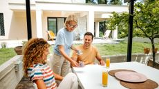 An older mother serves coffee to her adult daughter and son on a porch. A luxury home is in the background.