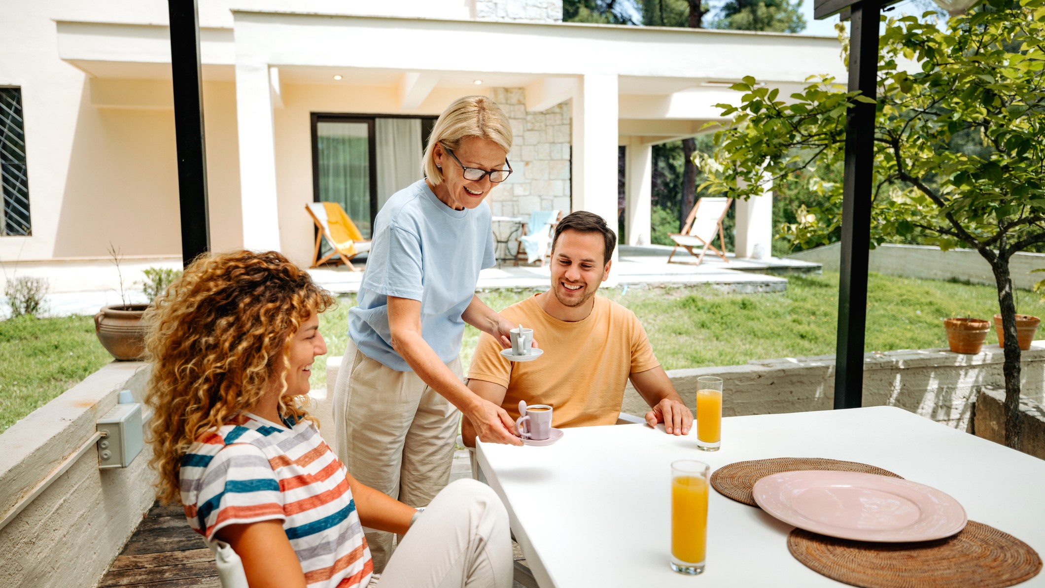 An older mother serves coffee to her adult daughter and son on a porch. A luxury home is in the background.