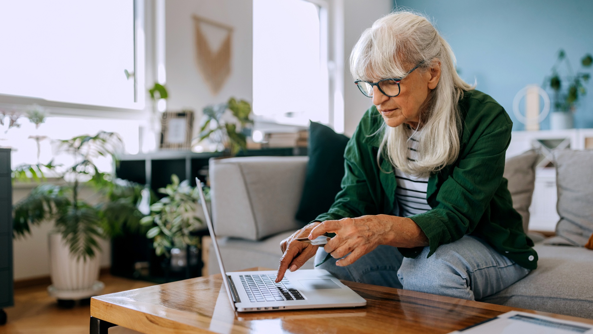 An older woman sits on her sofa and uses her laptop, which is sitting on her coffee table.