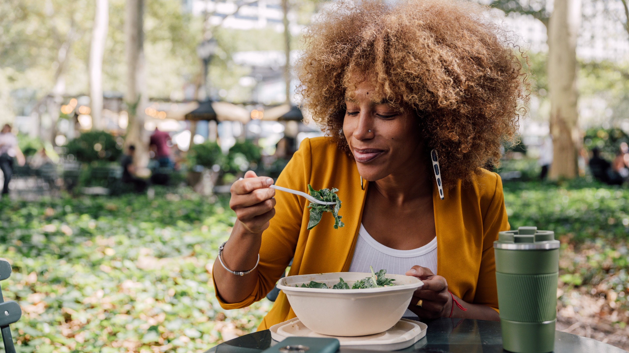 A woman in a park eats salad from a takeaway bowl.