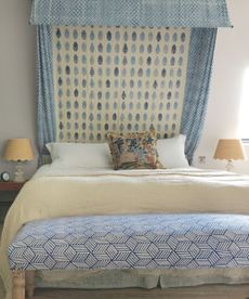 A blue bedroom with a patterned bed canopy and ottoman, matching beige scalloped lamps either side of the bed with white pillows, beige blanket and patterned cushion