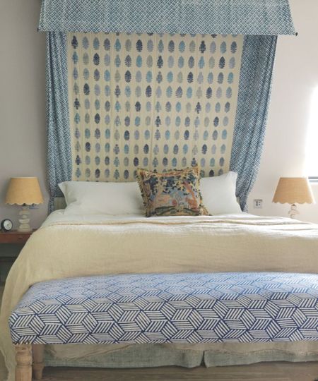 A blue bedroom with a patterned bed canopy and ottoman, matching beige scalloped lamps either side of the bed with white pillows, beige blanket and patterned cushion