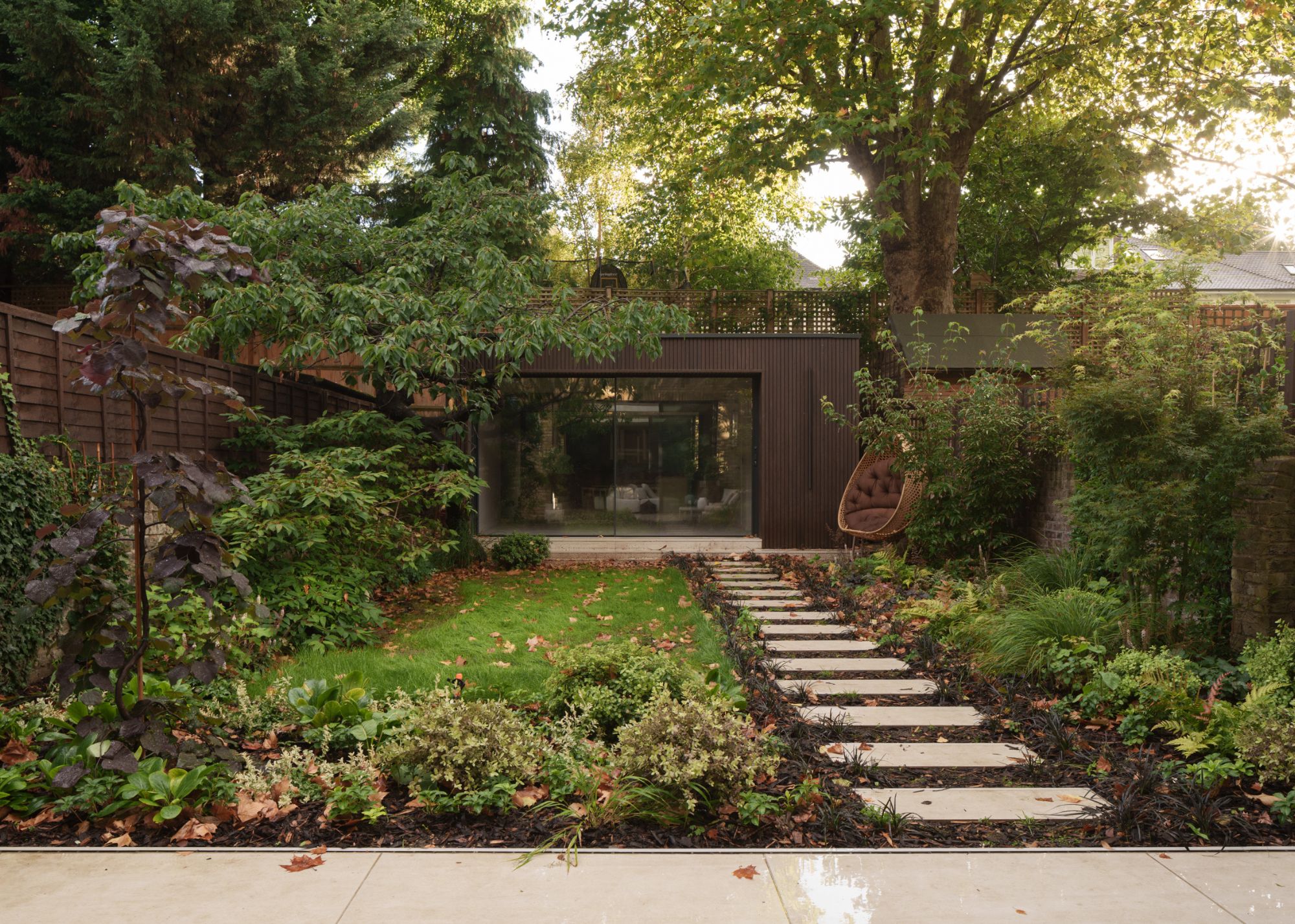 A natural-feeling garden with a spaces-slab pathway to a yoga room surrounded by plants and foliage with an egg chair at the far end of the space by the yoga room
