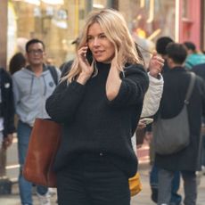 Sienna Miller wears cowboy boots, a black sweater, black jeans, and a brown suede tote bag.