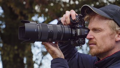 Man holding the Nikon Z8 camera up to his eye and the Nikon Z 70-200mm f/2.8 VR S II lens is attached, there are trees in the background