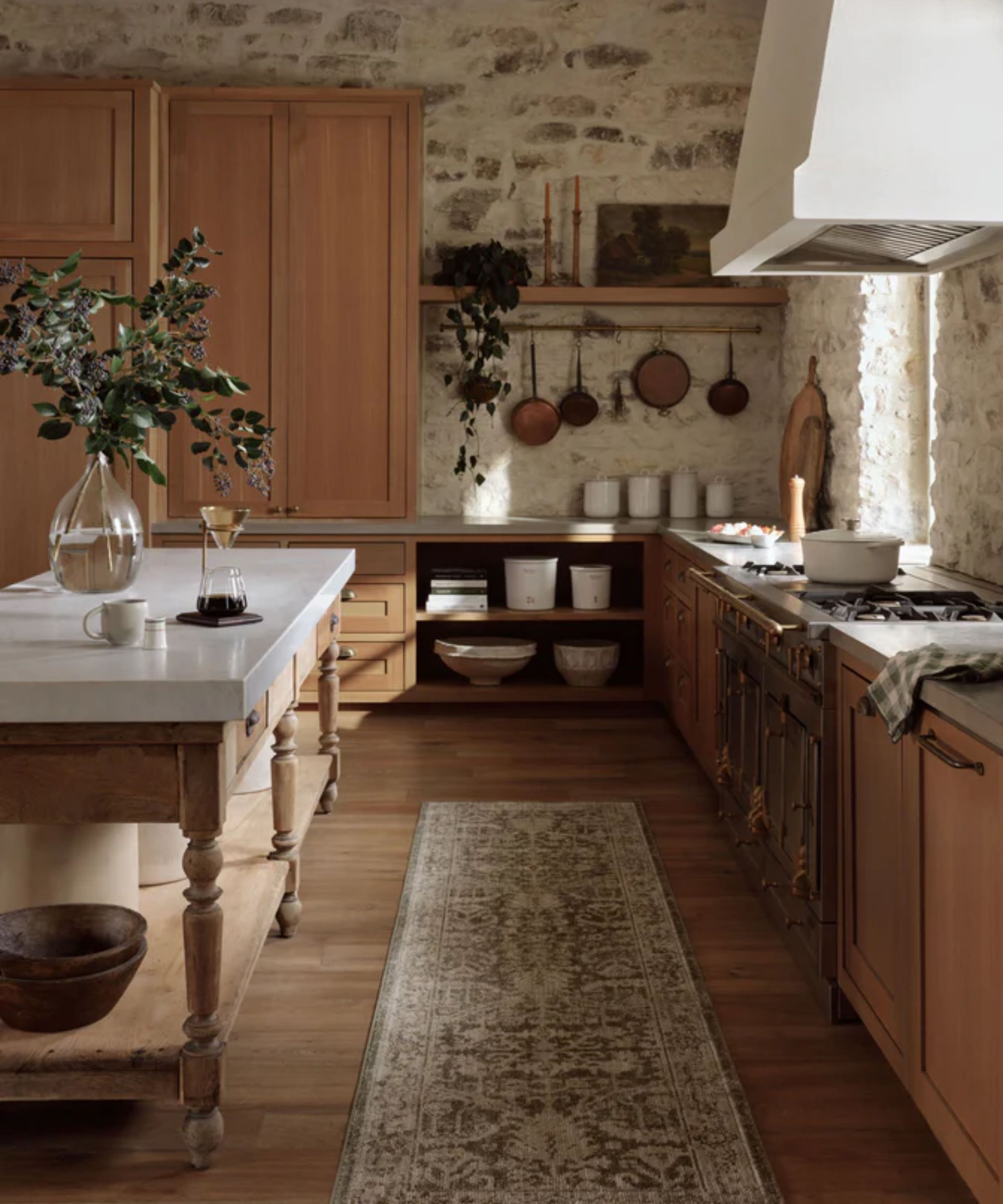 farmhouse kitchen with exposed stone walls, oak cabinetry, a plaster hood, wooden work table island with marble countertop top and a vintage style runner rug