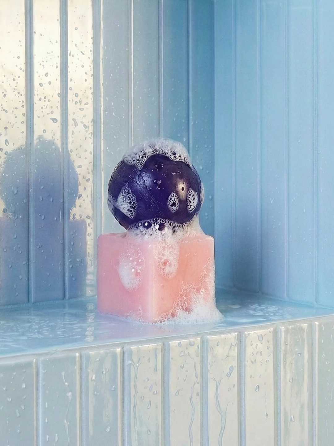 A pale pink square holder and a purple spherical soap in a blue tiled bathroom nook