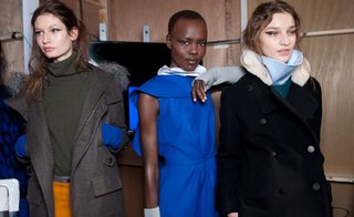 Three women, one with a brown coat over a brown jumper, one with a blue dress and white collar, and one with a black trench coat