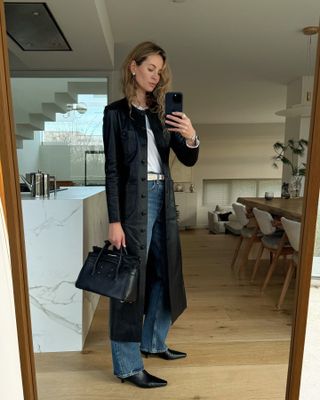 Woman taking mirror selfie wearing black collarless coat, black bag, blue jeans, and white shirt.