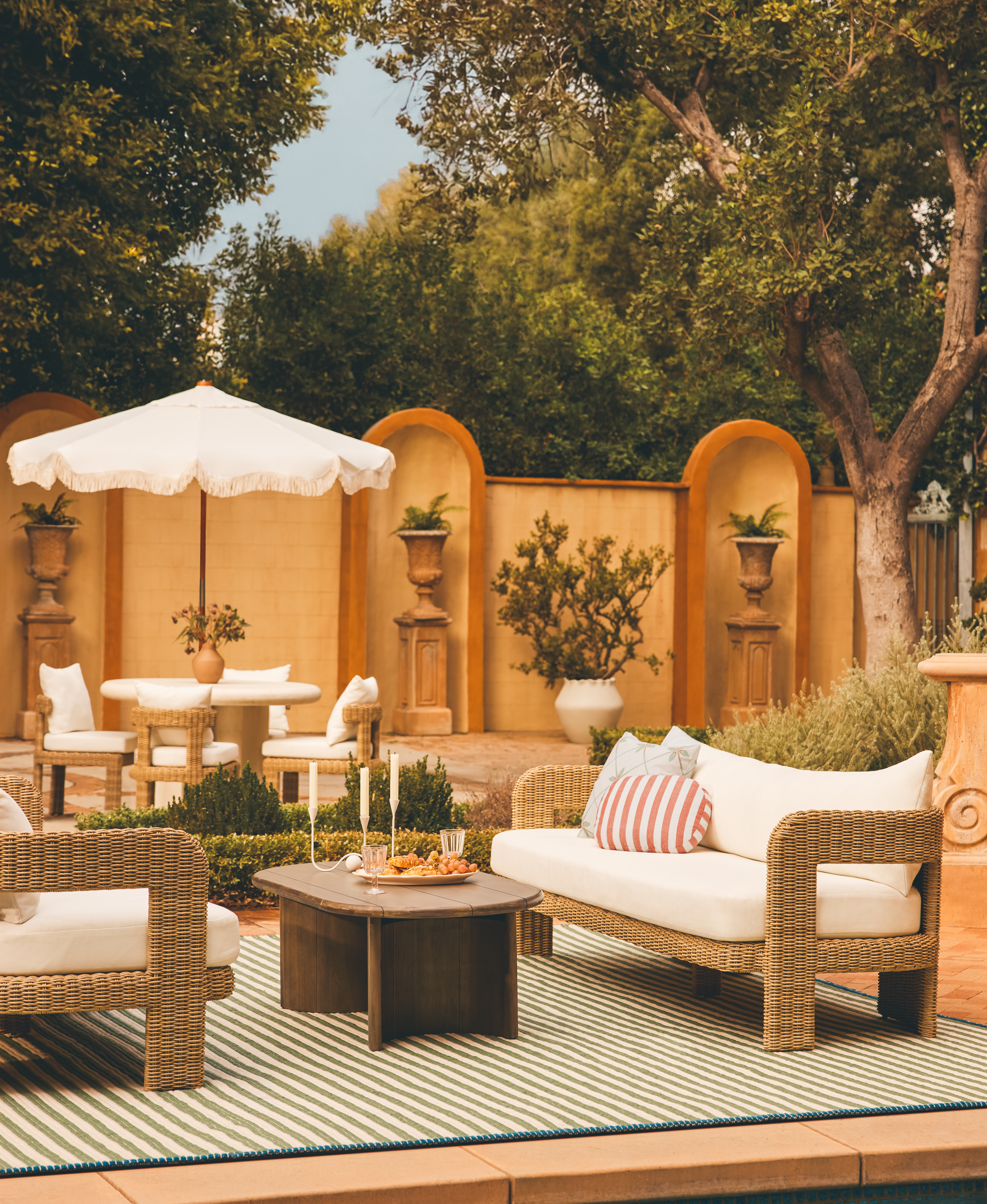 An outdoor seating area with a dining table and umbrella behind. There are decorative plants behind, and a wooden coffee table in between the sofas.