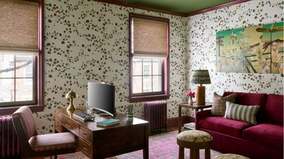 Home office with floral wallpaper, green ceiling, vintage wooden desk, burgundy velvet sofa, and a vibrant pink rug.