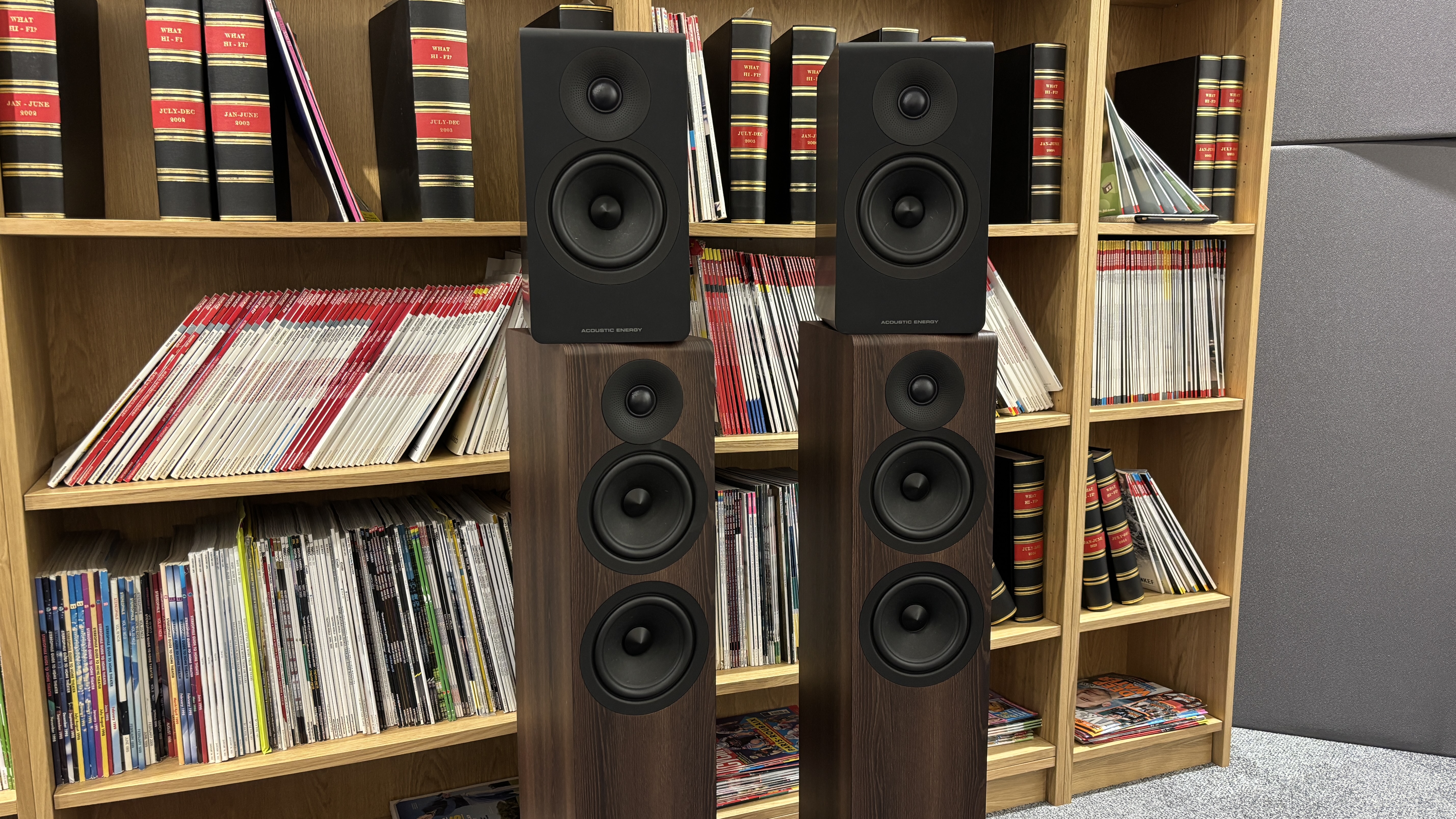 Acoustic Energy AE300 Mk2 sat on top of AE309 Mk2 next to each other in front of bookshelves filled with magazines