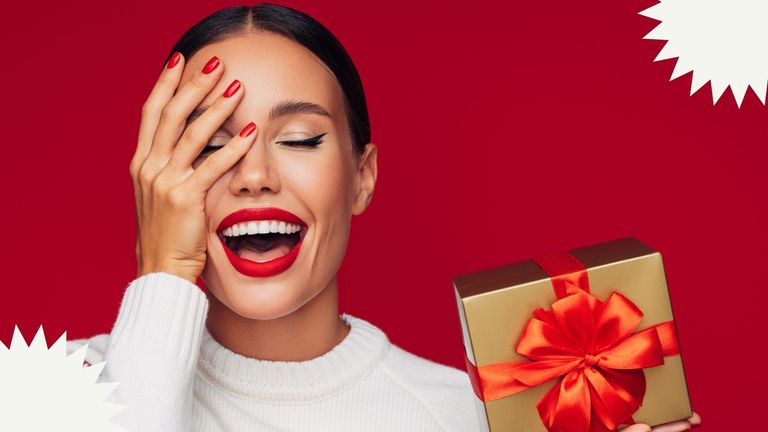 A woman wearing easy christmas makeup looks, a red lip and winged eyeliner holding a gift on a red backdrop